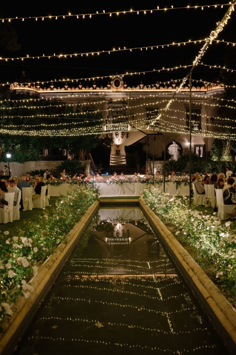 The top table at the head of a long reflecting pool at night, lights overhead, Taormina Weddings