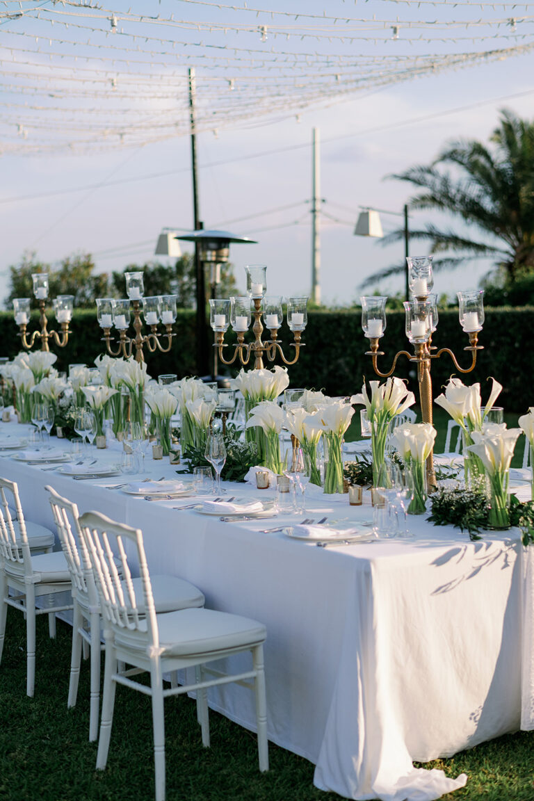 Brass candelabra and white calla lilies down a table under strung lights, Taormina Weddings