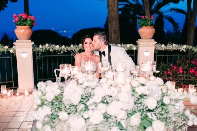 The couple at a table banked with white roses at blue hour, the coast lights behind, Taormina Weddings
