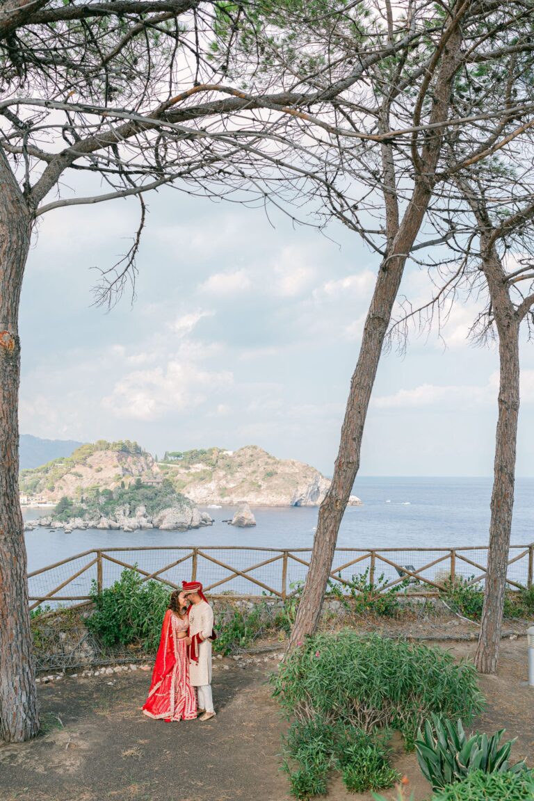 The couple in red and gold under stone pines, Isola Bella behind, Taormina Weddings