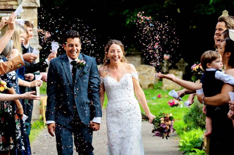 A bride in an off-the-shoulder lace gown and her groom walking through a shower of confetti, TDR Bridal