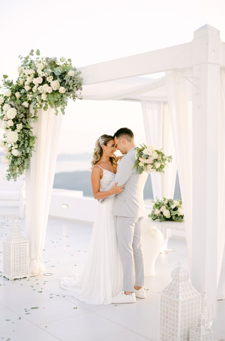 A couple kissing under a white canopy with a floral arrangement above a sea view, TDR Bridal
