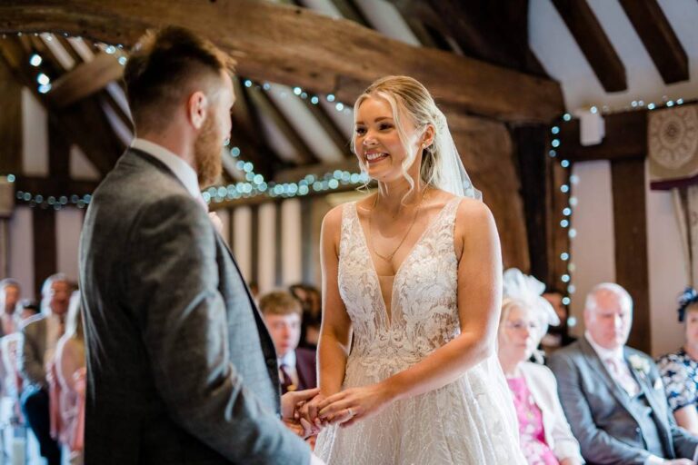 A bride and groom exchanging vows in a timber-framed barn with guests behind, TDR Bridal