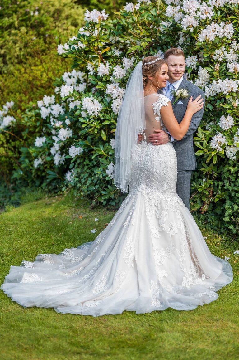 A bride in a lace mermaid gown with a long veil embracing her groom among flowering shrubs, TDR Bridal