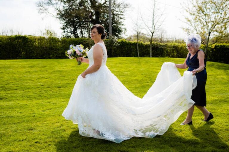 A bride in a lace gown on a sunlit lawn while a guest arranges her long train, TDR Bridal