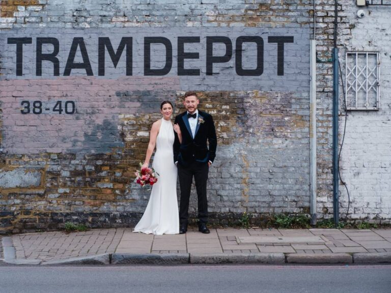 A bride and groom standing in front of a painted brick wall, the bride holding a bouquet, TDR Bridal