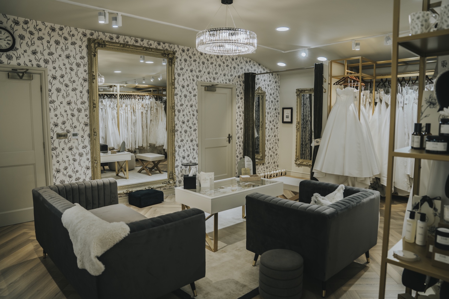 A lounge area with grey sofas, a gilt mirror and rails of white wedding gowns beneath a crystal chandelier, TDR Bridal