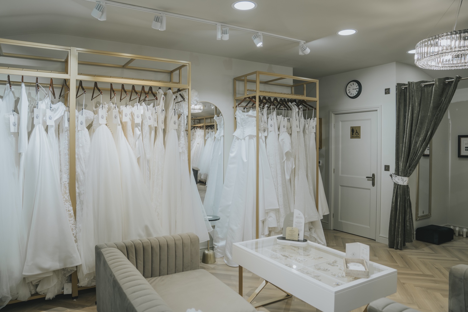Rails of ivory and white gowns beside a low white coffee table and pale seating, TDR Bridal