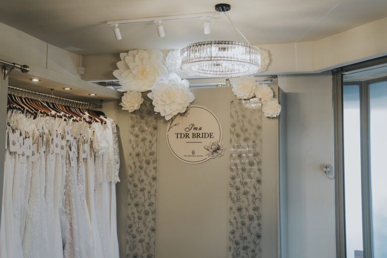 A wall of white gowns beside a circular I am a TDR Bride sign and paper flowers, TDR Bridal