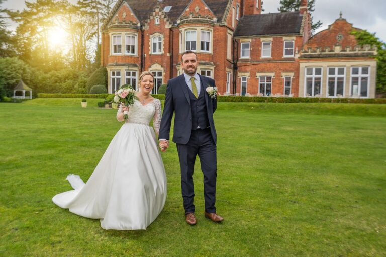 A bride in a long white gown walking hand in hand with her groom on a lawn in front of a red brick country house at sunset, TDR Bridal