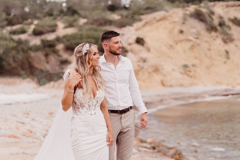 A bride in a lace gown with a floral headpiece and her groom walking on a sandy beach, TDR Bridal