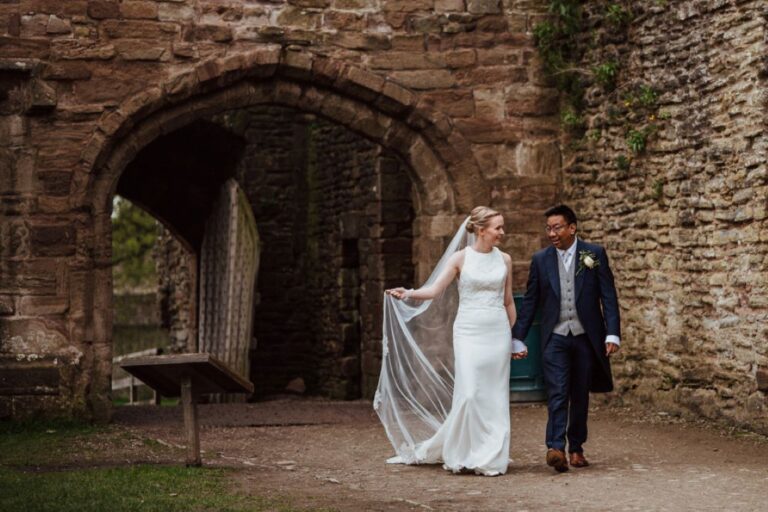 A bride holding her veil and walking with her groom beneath a stone ruin arch, TDR Bridal