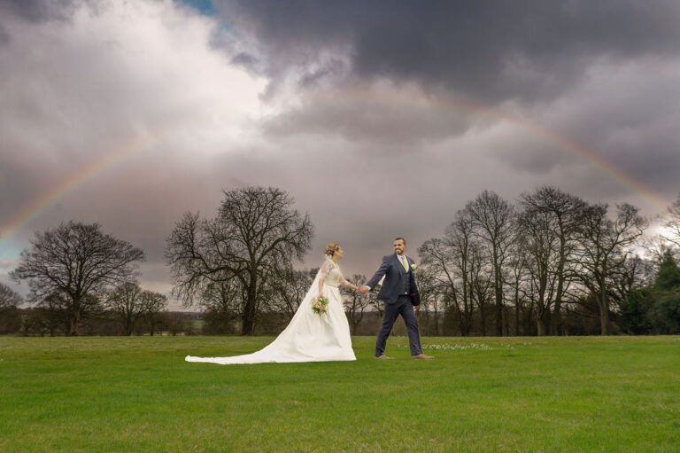 A bride and groom holding hands on parkland with a rainbow behind dark trees, TDR Bridal