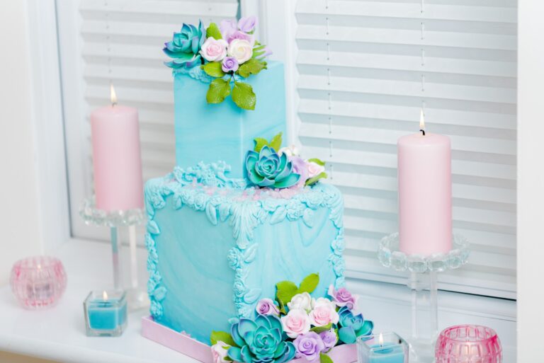 A square blue wedding cake with sugar succulents and flowers beside pink candles by a window, Tees Bakery