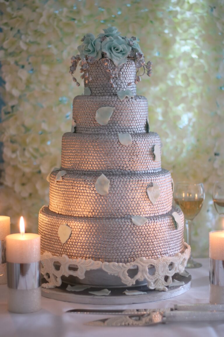 A tiered cake covered in silver beading with sugar leaves and a teal rose top, by candlelight, Tees Bakery