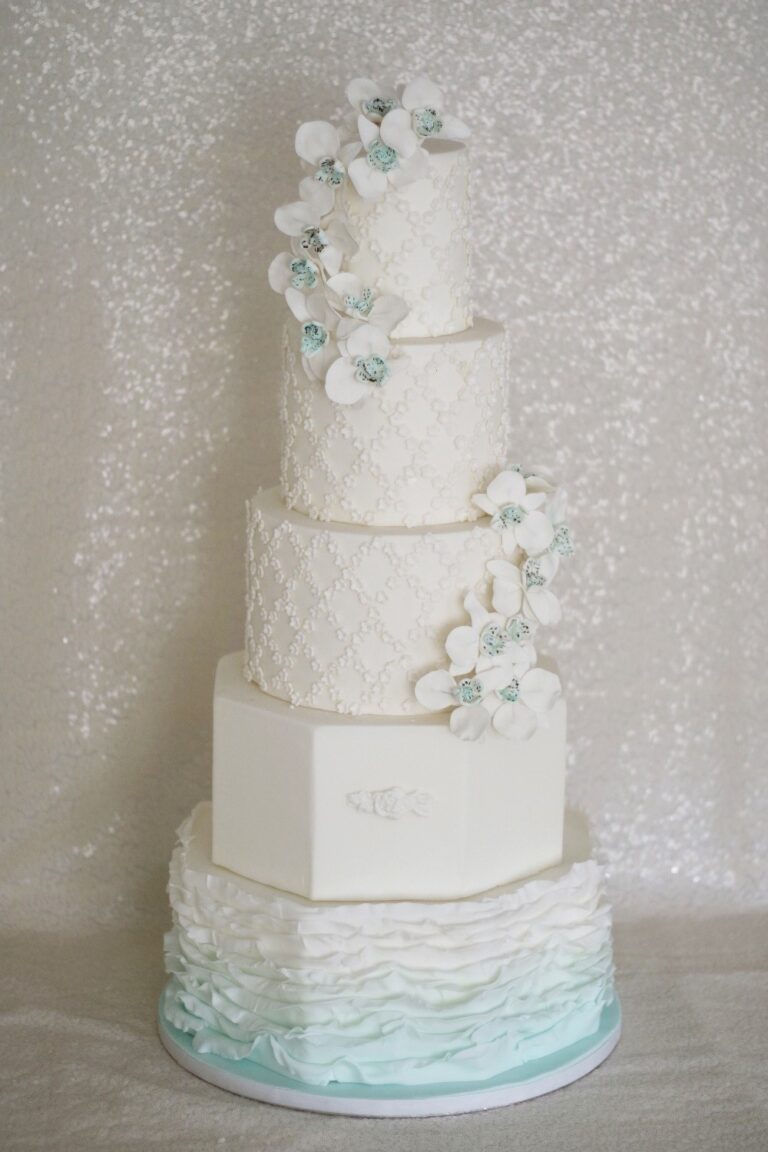 A tall white cake with lace pattern, blue ruffles and sugar orchids, Tees Bakery