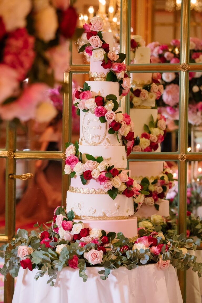 A tall white tiered cake wrapped in pink and red sugar roses and foliage, Tees Bakery