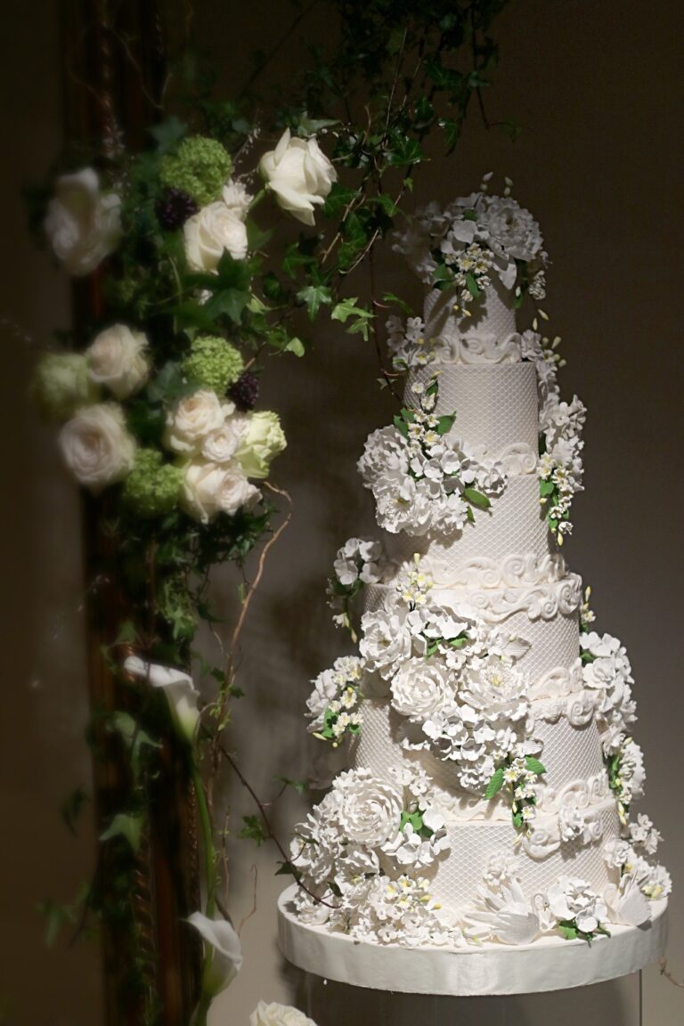 A tall white cake covered in sugar roses and blossom among fresh white roses and ivy, Tees Bakery