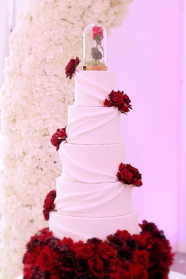 A white draped cake with red sugar roses and a glass dome topper above a base of red roses, Tees Bakery