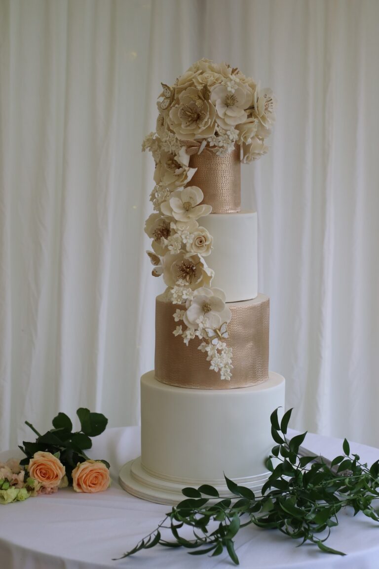 A white and champagne cake with a cascade of cream sugar flowers beside greenery, Tees Bakery