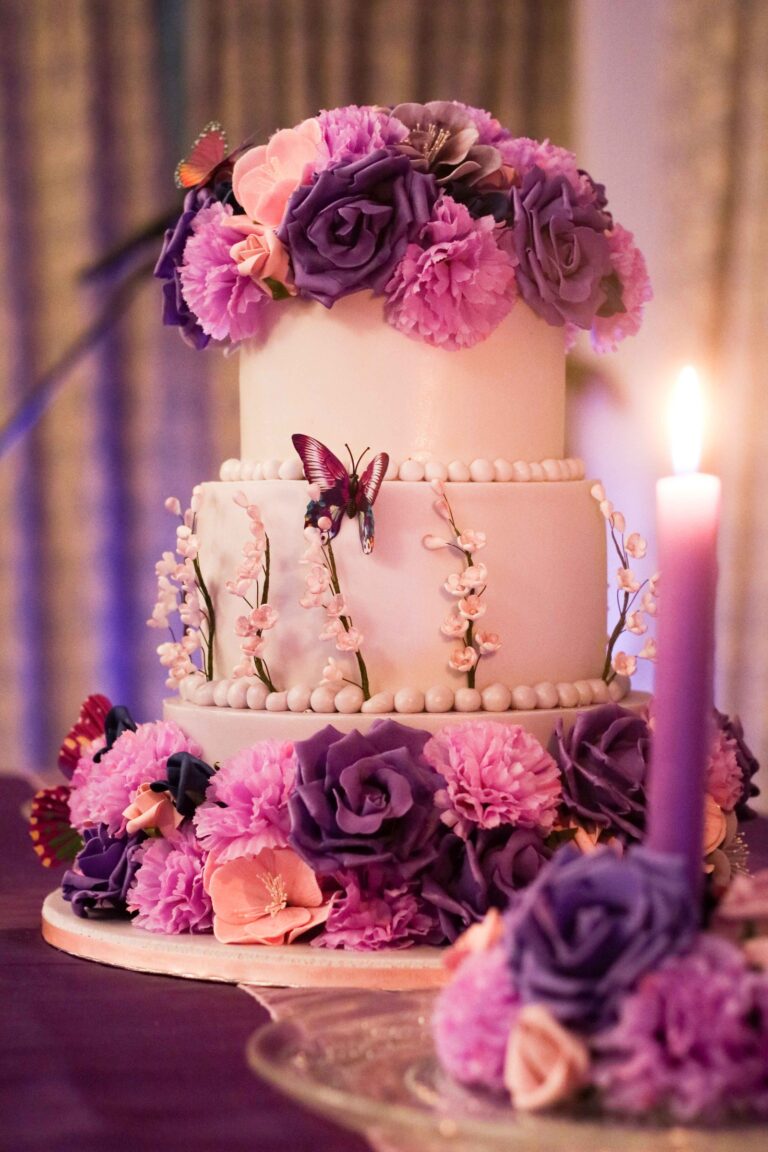 A tiered cake with deep purple and pink flowers, butterflies and a lit candle, Tees Bakery