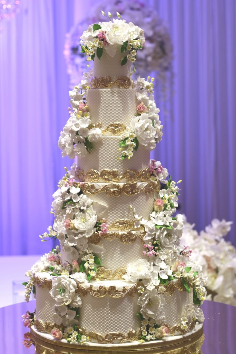 A tall hexagonal cake with gold piping and cream sugar flowers against lilac drapes, Tees Bakery