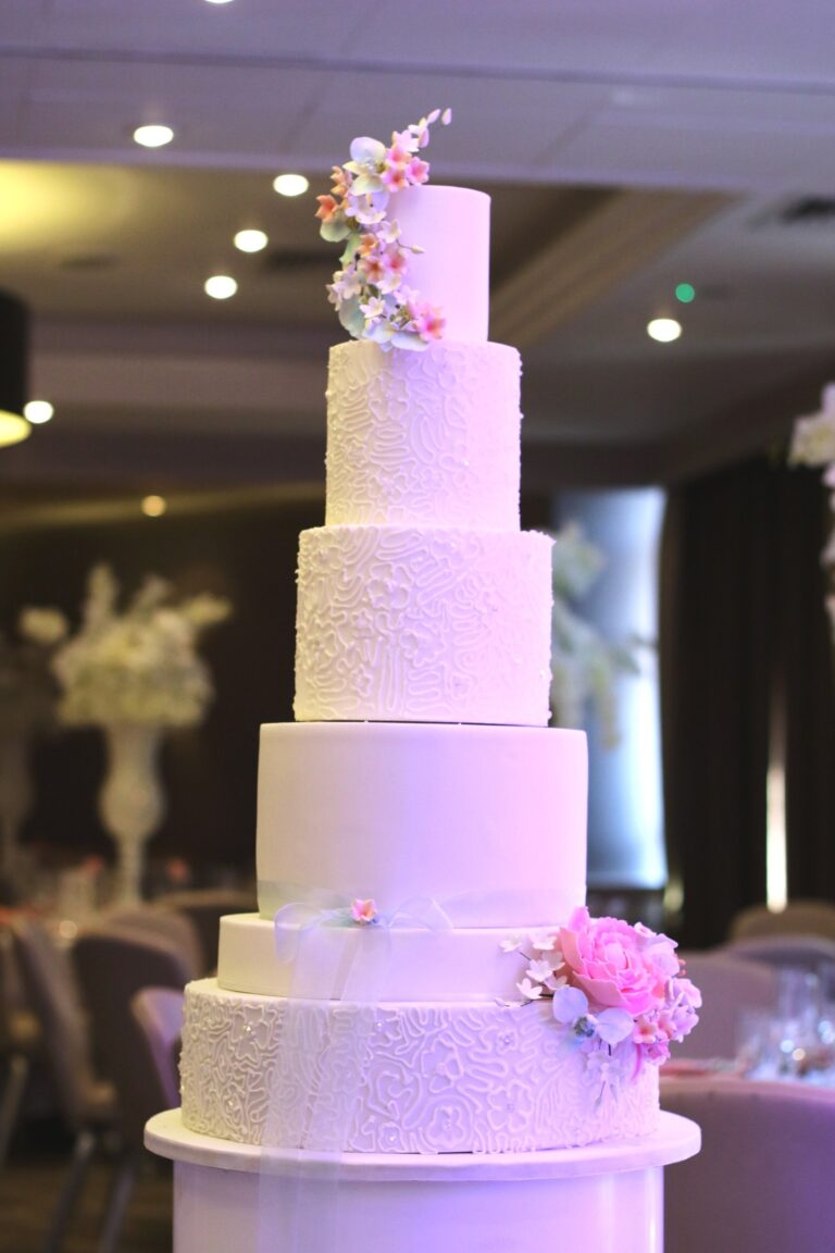 A five tier white cake with lace texture and a pink sugar flower on top, Tees Bakery