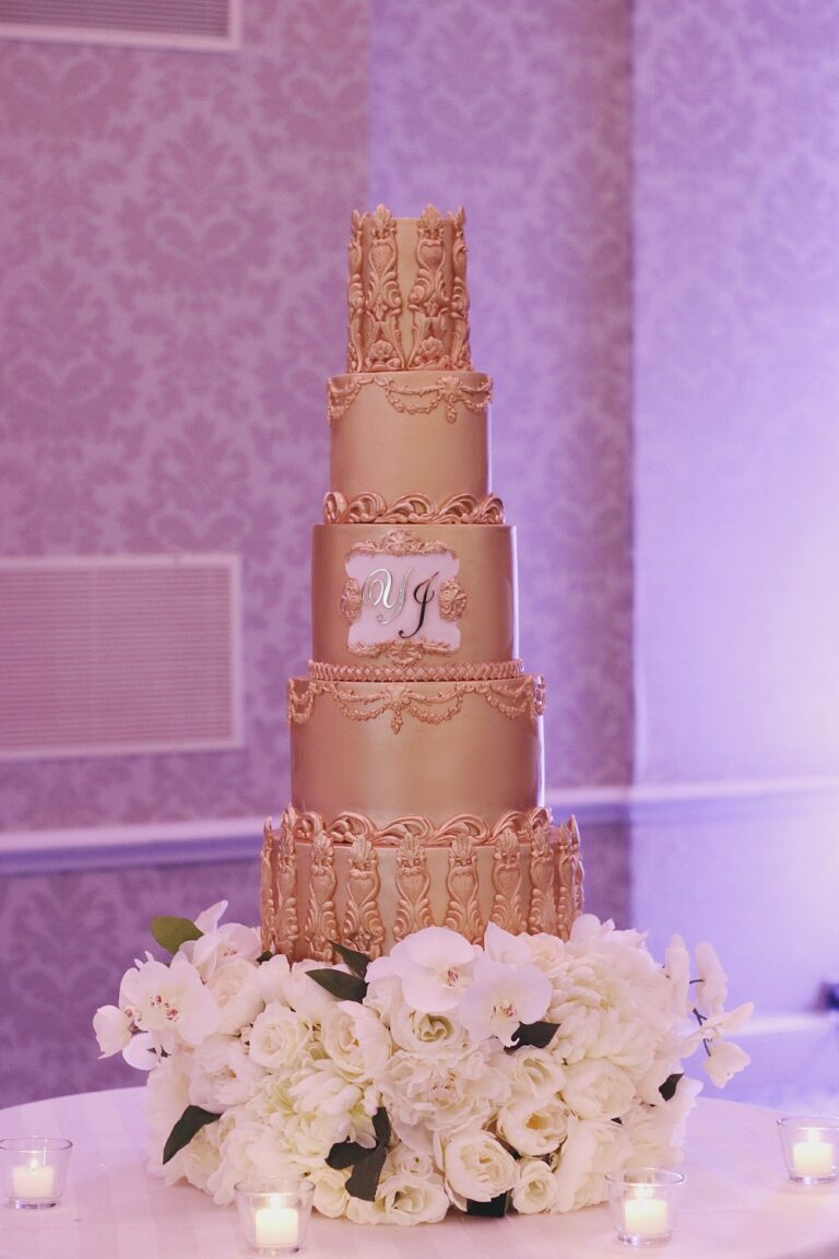 A rose gold and cream cake with a crest top and monogram panel on a bed of white roses, Tees Bakery