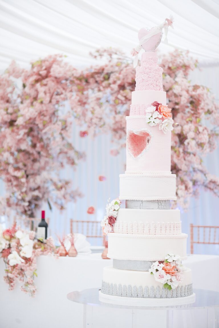 A tall white and pink cake with a heart panel before cherry blossom in a marquee, Tees Bakery
