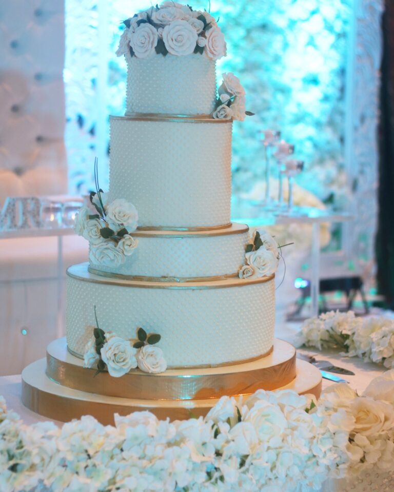 A pale blue and gold tiered cake with white sugar roses on a bed of white flowers, Tees Bakery