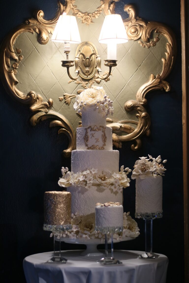 A tiered white cake with smaller satellite cakes on stands before a gilded wall light, Tees Bakery
