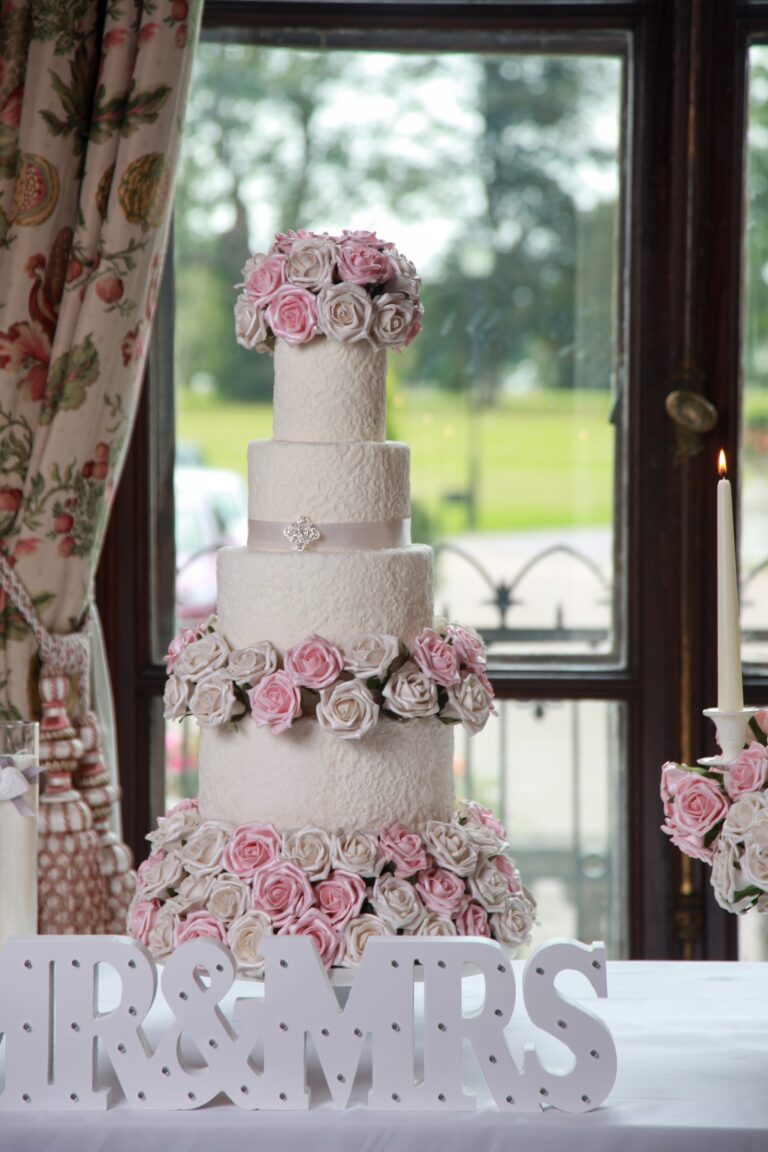 A white tiered cake with bands of pink sugar roses beside a Mrs and Mrs sign at a window, Tees Bakery