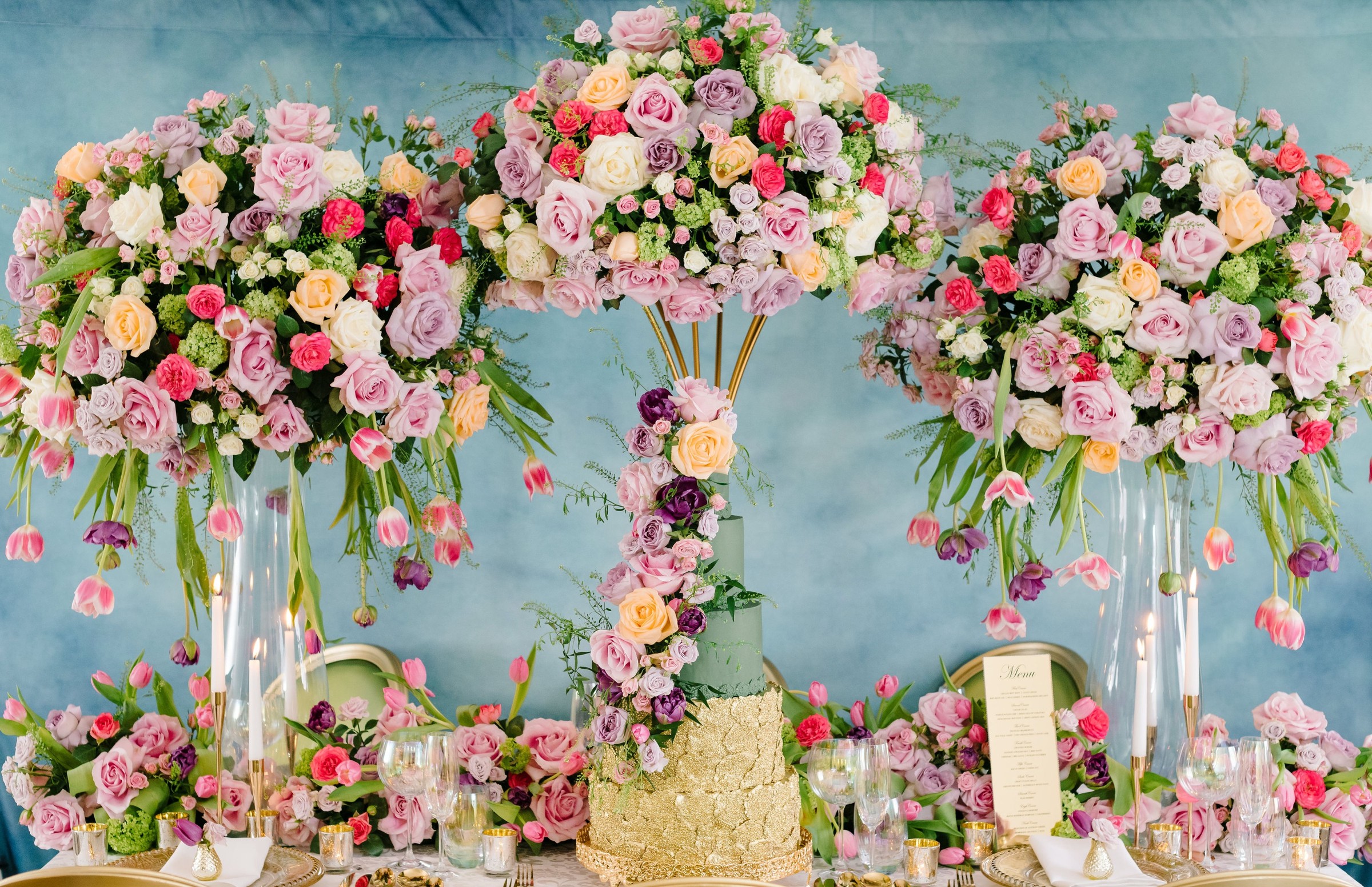 A gold wedding cake at the centre of a dessert table dressed with tall arrangements of roses and blossom, Tees Bakery