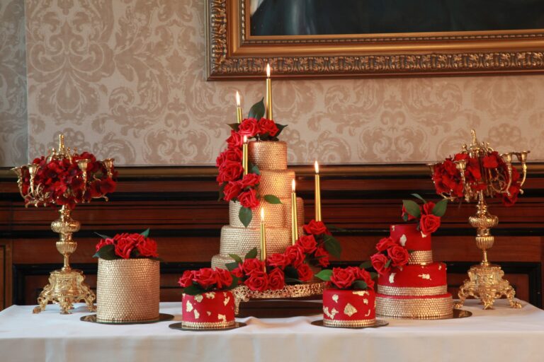 A tiered cake and smaller cakes in red and gold with candelabra on a dessert table, Tees Bakery