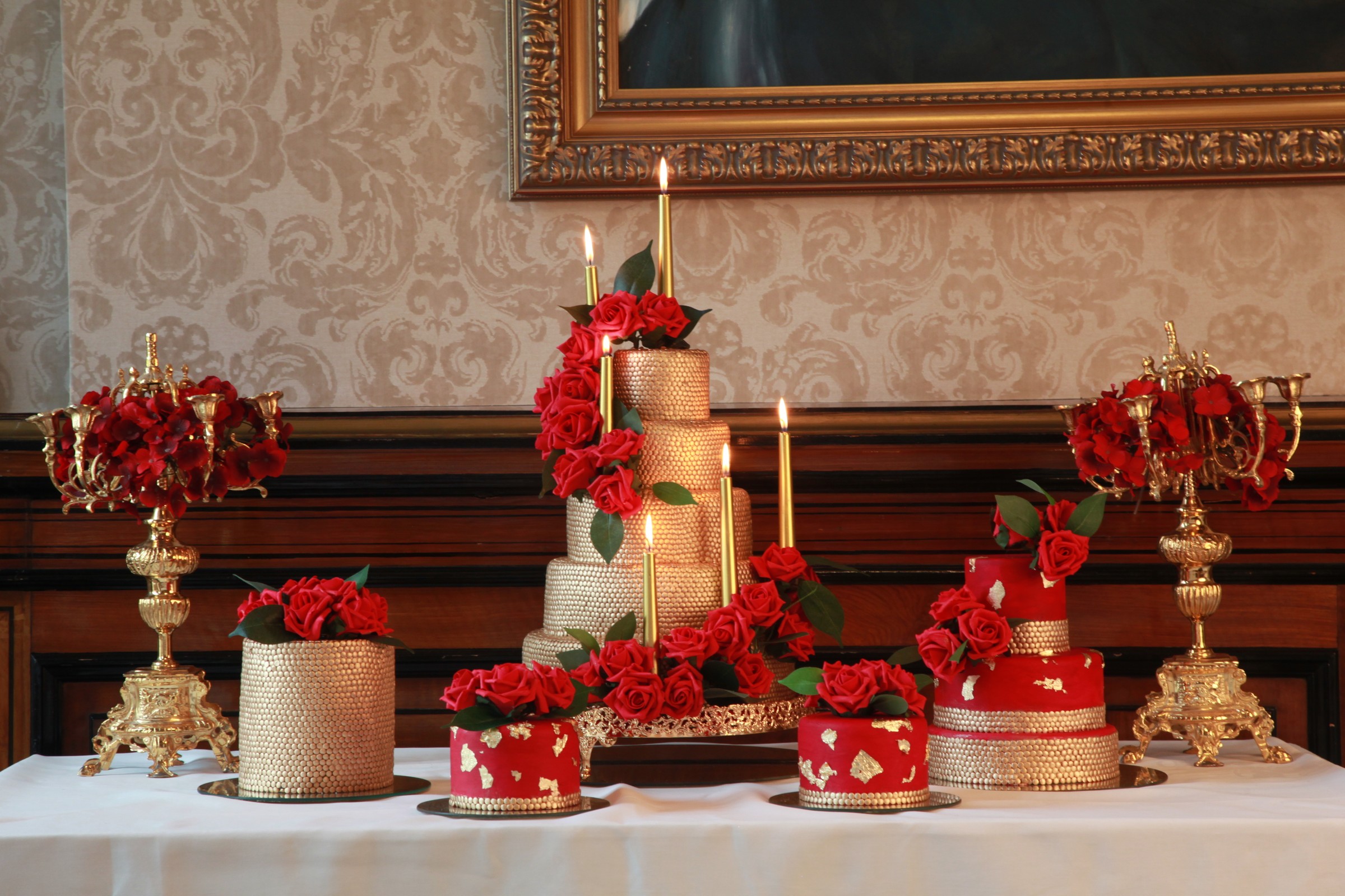 A tiered cake and smaller cakes in red and gold with candelabra on a dessert table, Tees Bakery