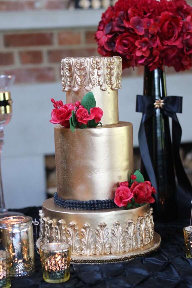 A gold and navy cake with red sugar roses and ornate gold piping on dark linen, Tees Bakery
