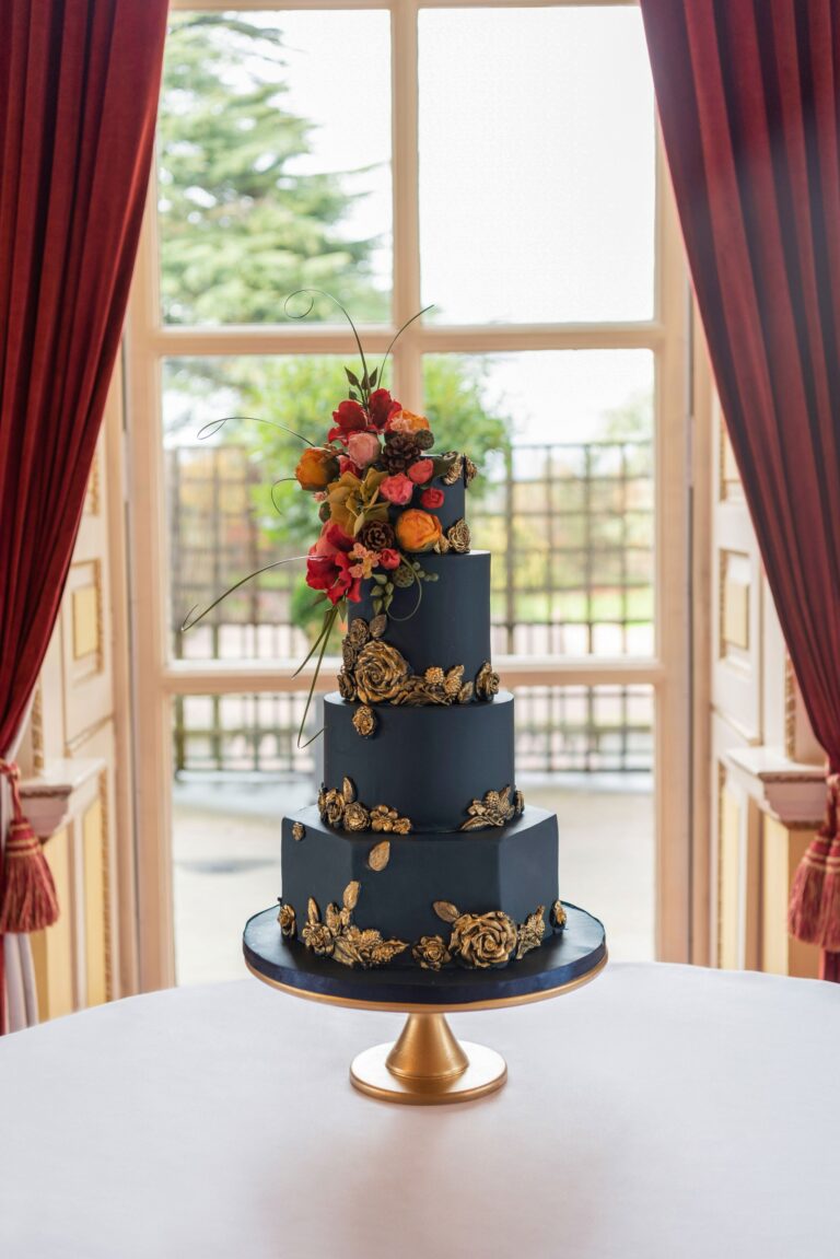 A navy tiered cake with gold detail and autumn flowers on a stand before a tall window, Tees Bakery