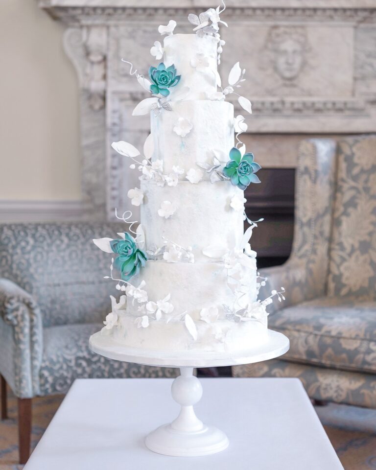 A tall white cake with sugar leaves and teal flowers beside a fireplace, Tees Bakery