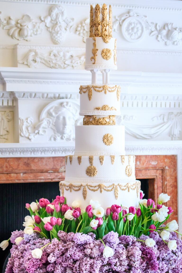 A white and gold cake with a crest topper above a bed of tulips and lilac before a marble fireplace, Tees Bakery