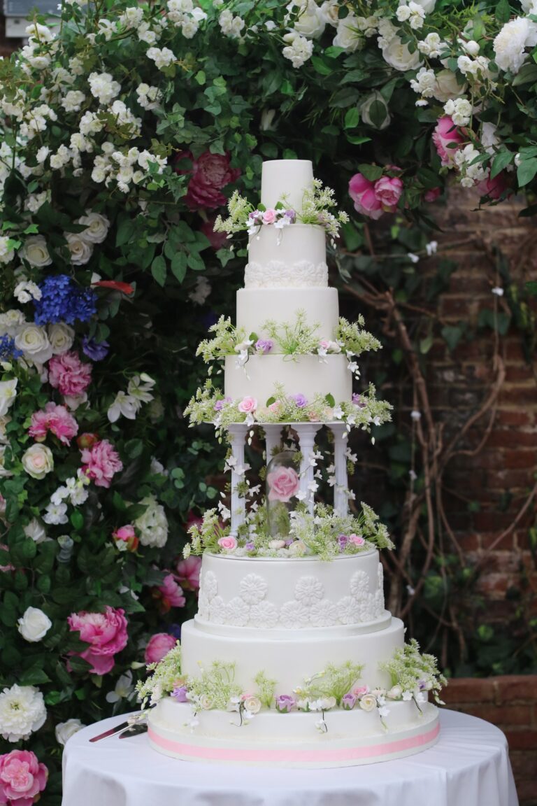 A white tiered cake raised on pillars before a flowering garden wall, Tees Bakery