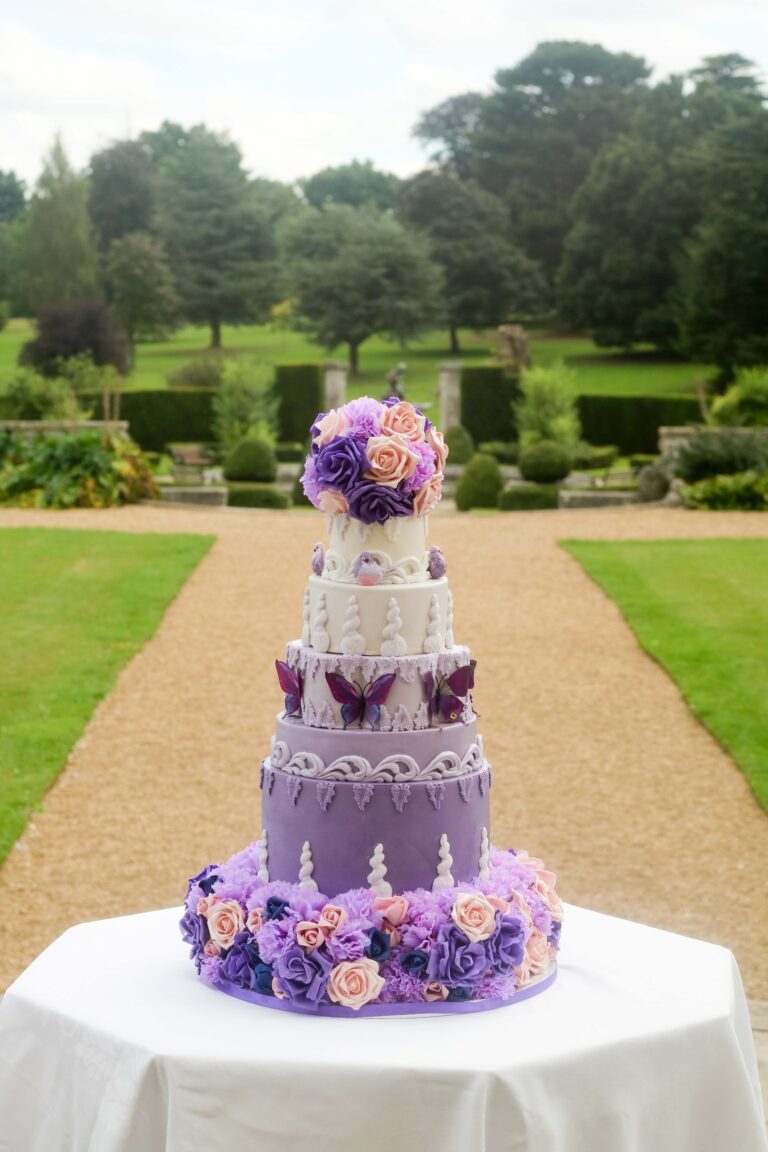 A purple and lilac tiered cake with sugar flowers at the end of a gravel path in a formal garden, Tees Bakery