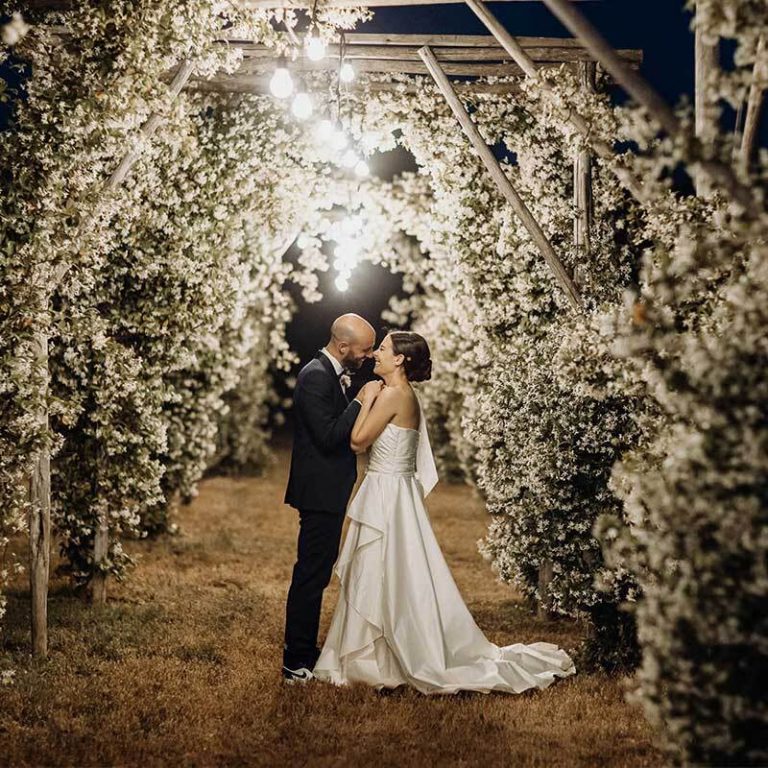 A couple beneath a pergola covered in white flowers and lit with string lights at night