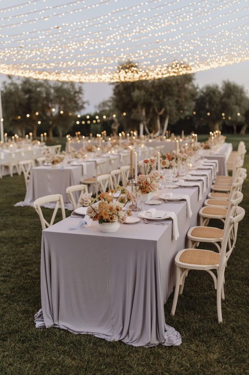 A long wedding table with cane-backed chairs on a lawn beneath festoon lights
