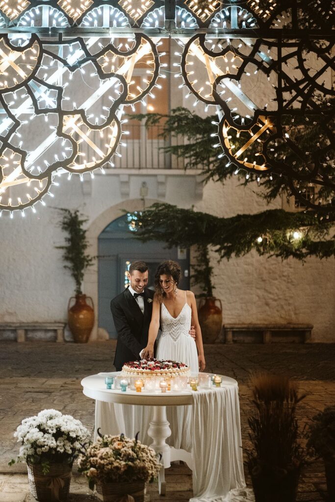 A couple cut their cake in the lit courtyard at night, Tenuta Pinto