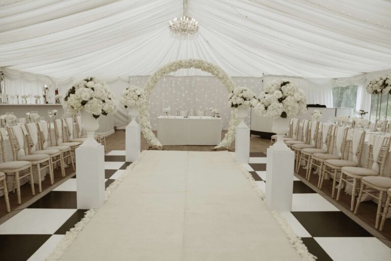 The marquee set for a ceremony, with a white aisle, chiavari chairs and a floral arch at the top table, That Amazing Place