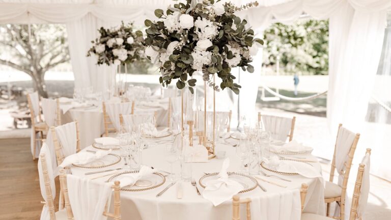A marquee table with gold-rimmed chargers, glassware and a tall white floral centrepiece, That Amazing Place