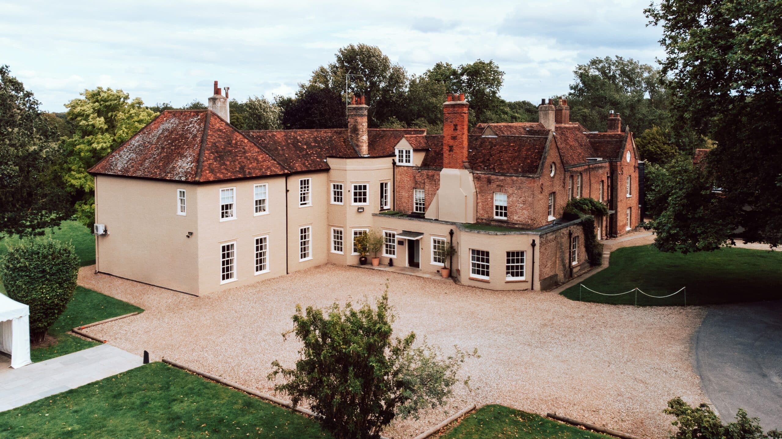 The front of the manor house from above the gravel forecourt: a cream rendered Georgian-style wing beside the older red brick range with tall chimneys, That Amazing Place
