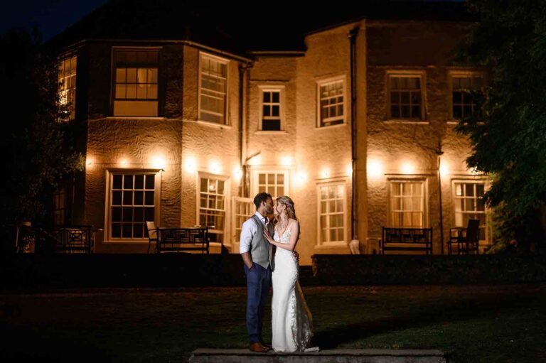 The rear terrace of the house lit up at night, with a couple on the lawn in front, That Amazing Place