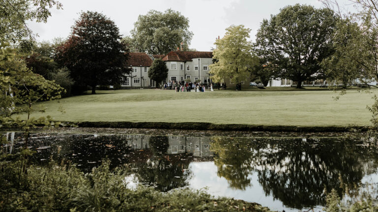 The house seen across the lake, with mature trees reflected in the water, That Amazing Place