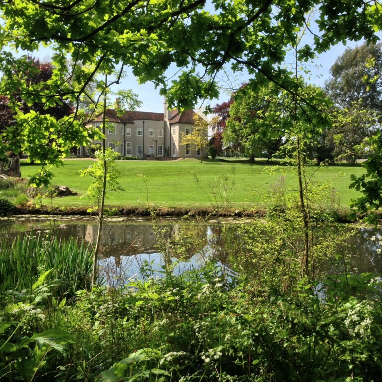 The lake in the foreground and the house beyond the lawn, framed by spring foliage, That Amazing Place